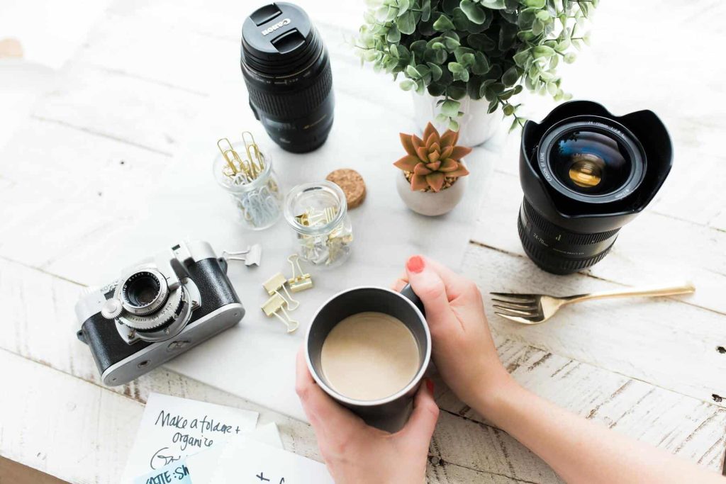 Top-down view of a photography desk with a camera, two lenses, jars of clips, a potted plant, and hands pouring coffee into a mug while notes about organizing photos are nearby.
