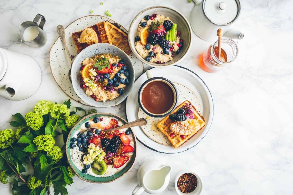 Overhead brunch spread with berry-topped oatmeal bowls, waffles, toast, and a teapot on a marble table.