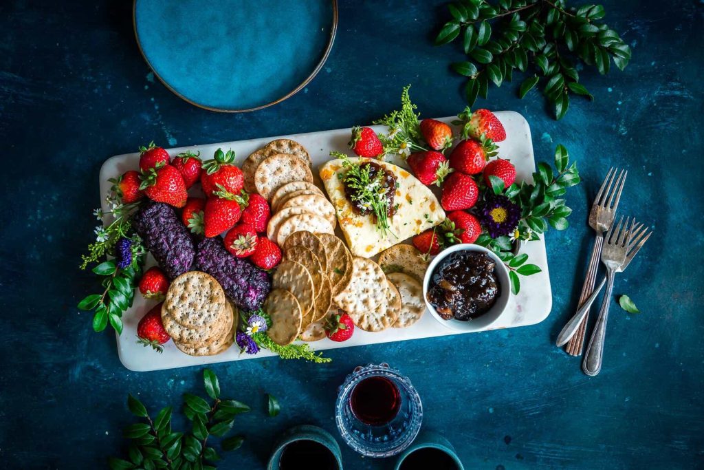 Charcuterie board on a teal table with crackers, cheese, strawberries, greens, and a small cup of jam; forks nearby.