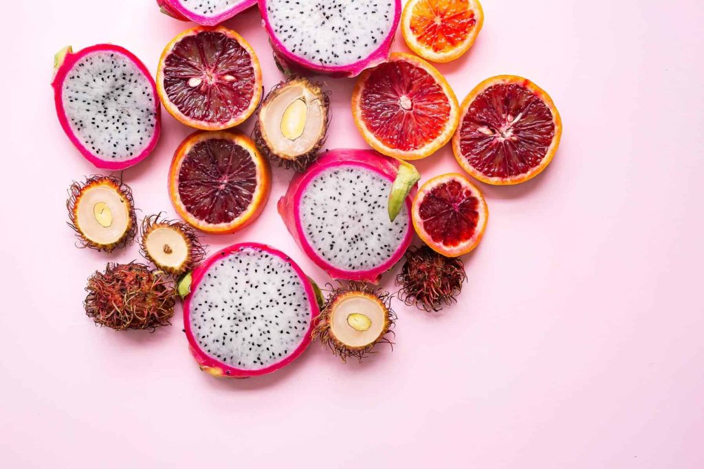 Assorted sliced tropical fruits scattered on a pink background, including dragon fruit, oranges, blood oranges, rambutan, and longan.