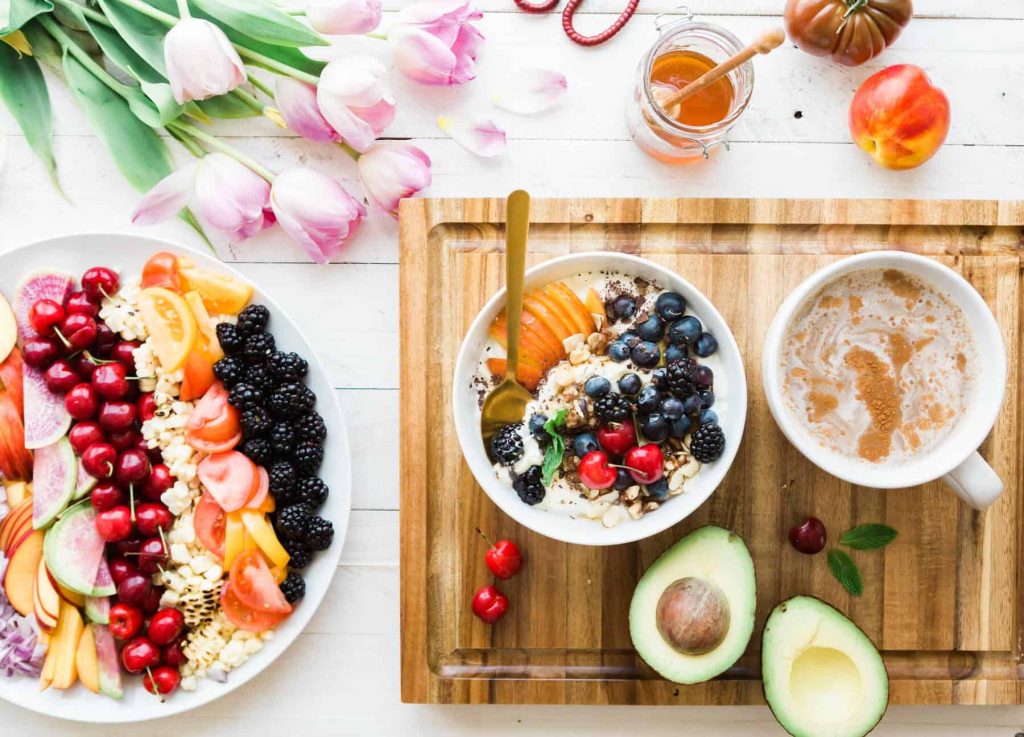 Top-down view of a breakfast spread: bowls of yogurt with granola and mixed berries alongside a fruit platter and avocado halves on a wooden board and white table.
