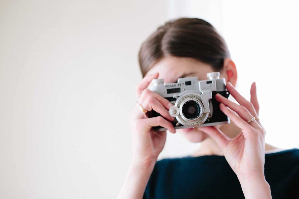 Woman holding a vintage silver camera up to her face, looking through the viewfinder.