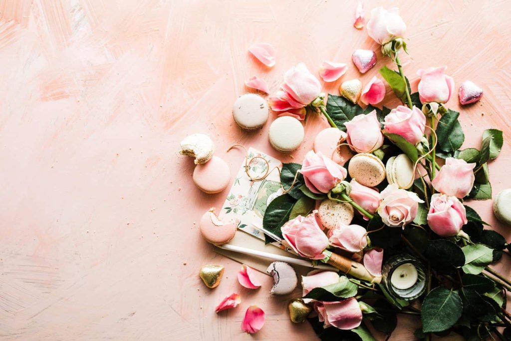 Flat lay of pink roses, macarons, and heart-shaped chocolates on a soft pink background close to a romantic dessert setup.