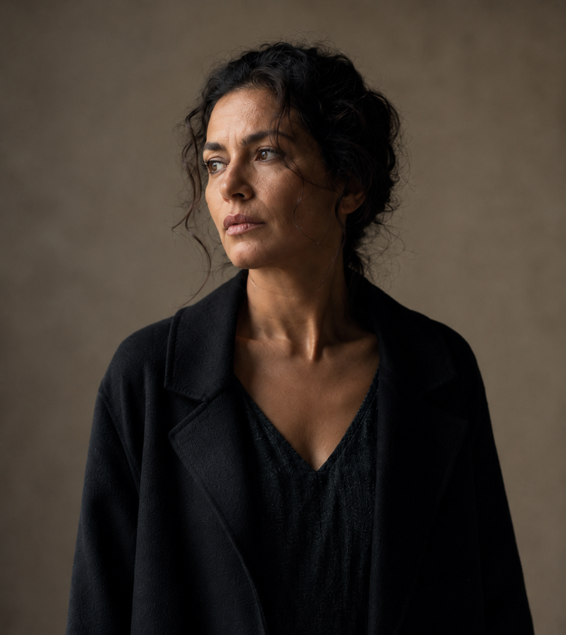 Portrait of a woman with dark hair wearing a black coat, looking off to the left against a neutral brown background.