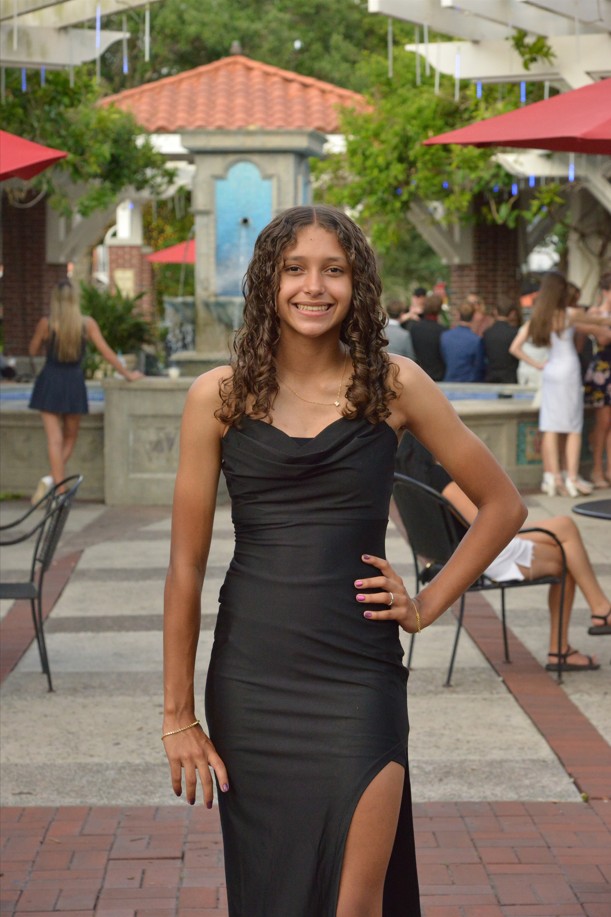 Young woman in a black evening gown posing with hand on hip at an outdoor event courtyard with a fountain in the background.